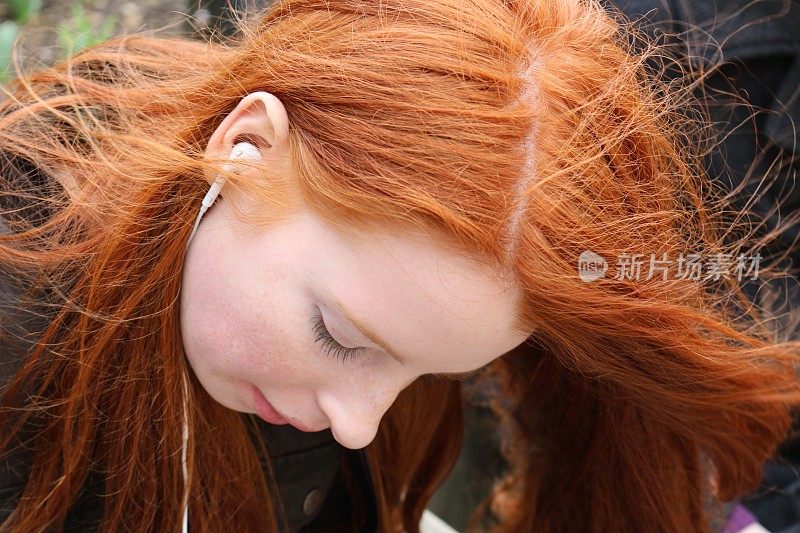 14 / 15岁的红发少女，皮肤苍白，脸上有雀斑，透过耳机听音乐，向下看照片